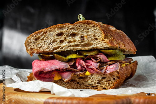 Pastrami sandwich on rye bread with pickles and mustard sauce, served on  wooden plate