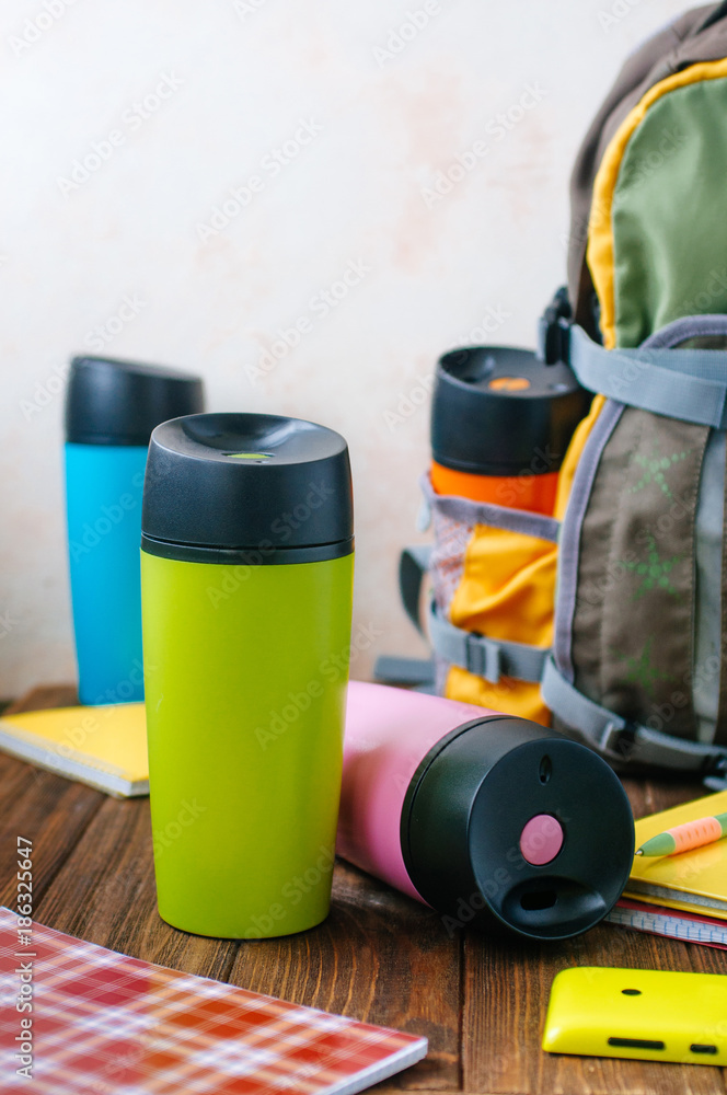 Color plastic thermos mugs near backpack on wooden background