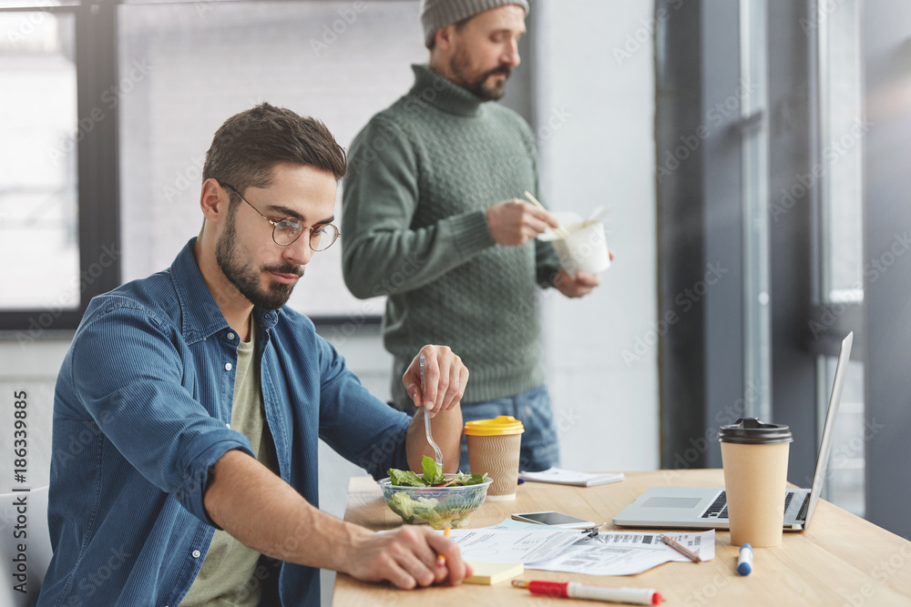Handsome brunet male enterpreneur being vegeterian, eats healthy vegetable salad at office, studies documents attentively and his partner stands in background, eats fast food with chopsticks Handsome brunet male enterpreneur being vegeterian, eats healthy vegetable salad at office, studies documents attentively and his partner stands in background, eats fast food with chopsticks
