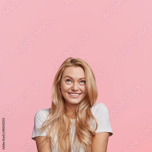 Glad blonde woman looks with cheerful shy expression upwards, smiles in charming way, has first date with handsome boyfriend, feels awkwardly, isolated over pink background. Positive human emotions