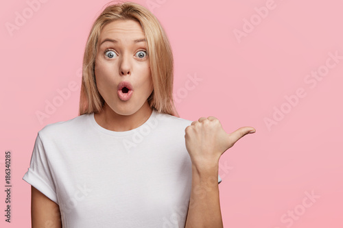 Horizontal shot of stupefied female model with surprised expression, keeps mouth opened, not expect to see something, indicates with thumb at blank studio wall for your advertisment or promotion
