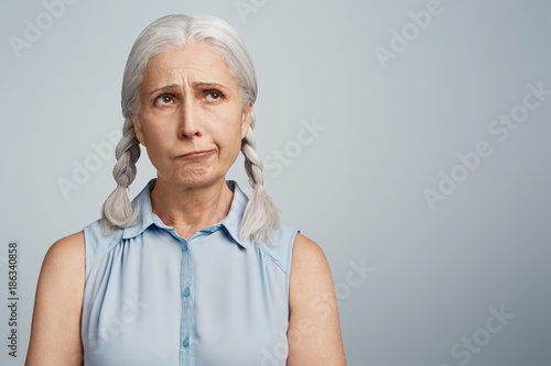 Thoughtful pensive hesitant mature woman with two grey pigtails, dressed in blue blouse, looks upwards, tries to male decision, isolated over blank studio wall with copy space for your hearder photo