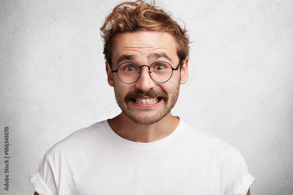 Funny attractive bearded young male with comic smile, has awkward look, pretends to be happy, looks gladfully through round spectacles, laughs at joke, isolated over white concrete background. Funny attractive bearded young male with comic smile, has awkward look, pretends to be happy, looks gladfully through round spectacles, laughs at joke, isolated over white concrete background.