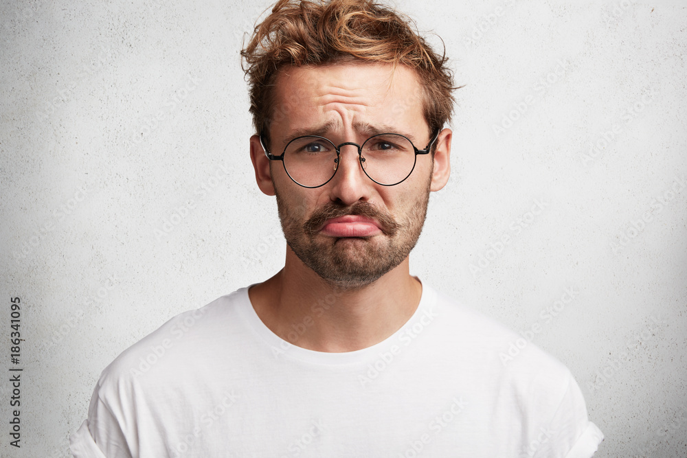 Depressed bearded male model curves lips and looks stressfuly at camera, being not ready to sustain defeat, has regretful expression, feels lonely and bored, isolated over white studio background Depressed bearded male model curves lips and looks stressfuly at camera, being not ready to sustain defeat, has regretful expression, feels lonely and bored, isolated over white studio background