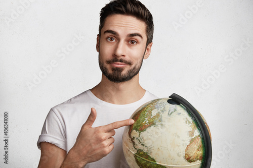 Portrait of serious confident unshaven man with trendy hairstyle, holds globe, chosses destination for travelling with girlfriend, likes to explore new unknown places, isolated over white background photo