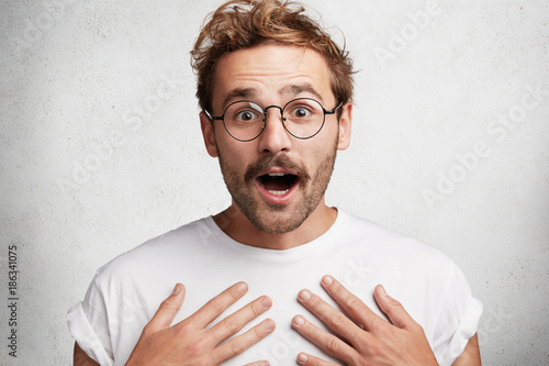 Foto Amazed young stylish guy keeps hands on chest, opens mouth in bewilderment, stares at camera, surprised to recieve big pecuniary reward, isolated over concrete studio background