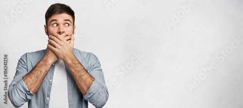 I will not say anything! Young male model has dark hair covers mouth with hands, dressed casually, isolated over white concrete wall with copy space for your text. Attractive man tries keep silence
