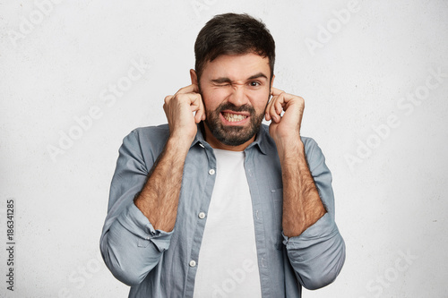 Foto Handsome bearded young male model plugs ears with displeased expression as hears annoying loud noise or roaring not far from him, isolated over white concrete wall