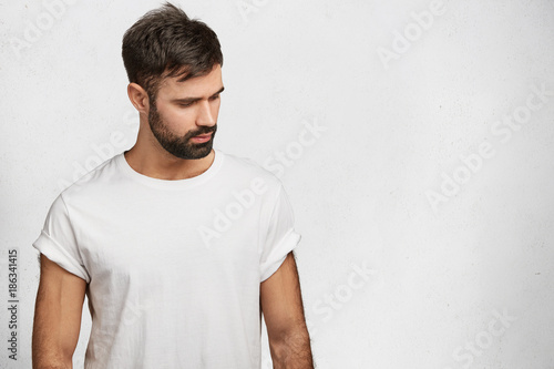 Sporty muscular man with trnedy hairdo, beard and mustache, wears white t shirt, looks with dreamy expression down, isolated over white concrete wall with copy space for your advrtisment or hearder photo