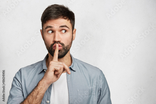 Serious good looking male with trendy hairdo, keeps fore finger on lips, tries to keep conspiracy, looks aside, says: Shh, make silence please. Isolated shot of attractive man shows silence sign photo