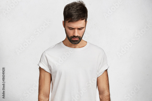 Handsome brunet male with trendy hairdo, has muscular body, wears caual white t shirt, looks down as notices something on floor, poses against concrete background. Attractive man hangs head.