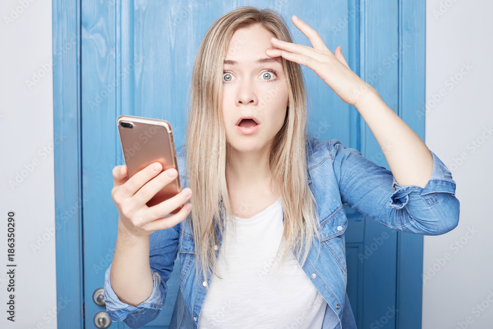 Headshot of young blond attractive woman with bugged eyes and widely opened mouth holding her hand on head staring at camera with shocked expression, amazement and great wonder. People and emotions. Headshot of young blond attractive woman with bugged eyes and widely opened mouth holding her hand on head staring at camera with shocked expression, amazement and great wonder. People and emotions.