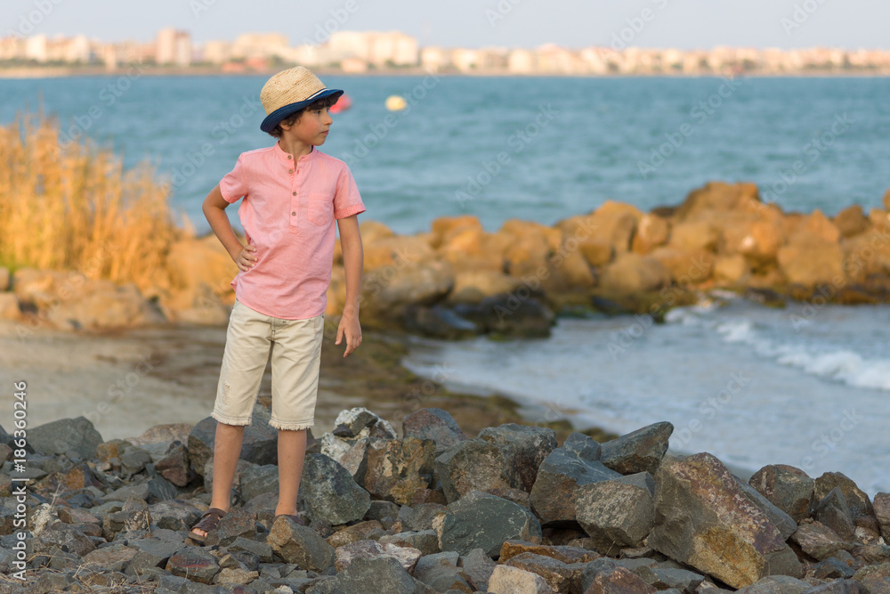 The boy stands on the stony seashore and looks into the distance.