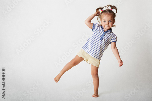 Принти на полотні Small adorable female child has funny expression, two pony tails, wears casaul t shirt and shorts, stands on one leg, poses against white background