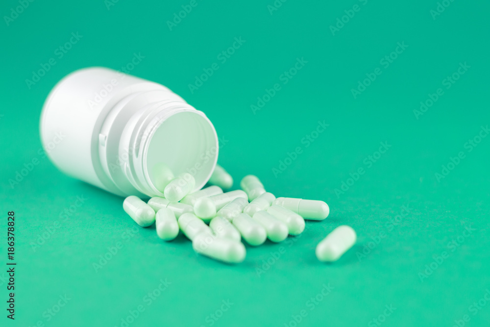 Close up white pill bottle with spilled out capsules on emerald green background with copy space. Focus on foreground, soft bokeh. Pharmacy drugstore concept Close up white pill bottle with spilled out capsules on emerald green background with copy space. Focus on foreground, soft bokeh. Pharmacy drugstore concept