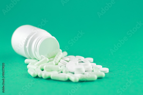 Close up white pill bottle with spilled out pills and capsules on emerald green background with copy space. Focus on foreground, soft bokeh. Pharmacy drugstore concept