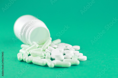Close up white pill bottle with spilled out pills and capsules on emerald green background with copy space. Focus on foreground, soft bokeh. Pharmacy drugstore concept