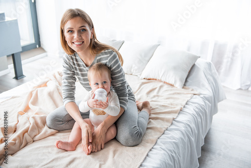 Portrait of smiling female and her little girl sitting on bed, kid is drinking milk from feeding bottle. Copy space in right side