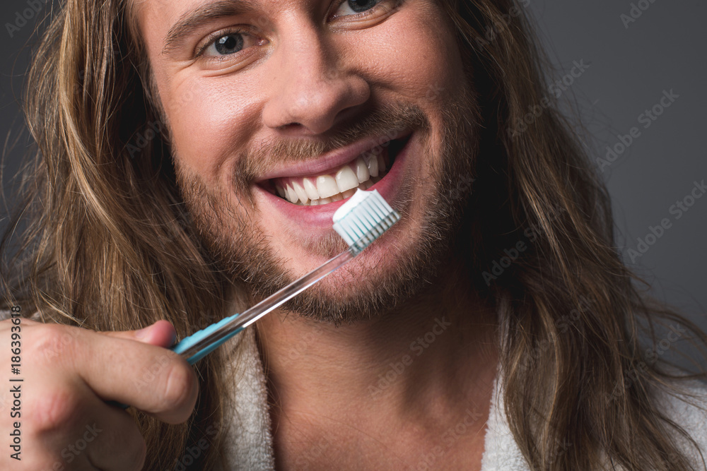 Personal hygiene. Close up of pleasant bearded man holding toothbrush and smiling. Isolated on grey background Personal hygiene. Close up of pleasant bearded man holding toothbrush and smiling. Isolated on grey background