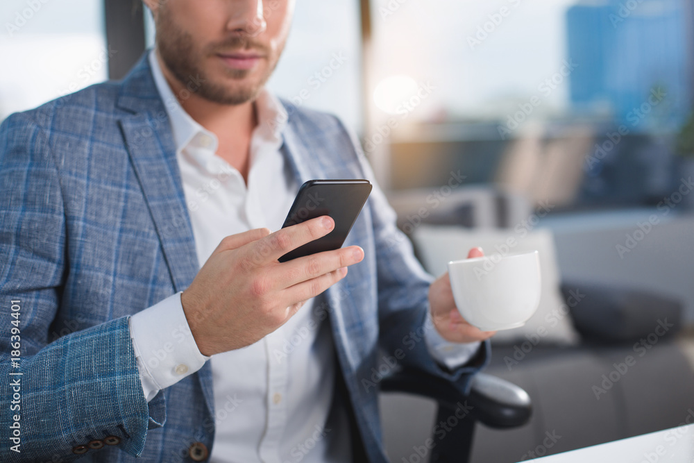 Intelligent male taking break after hard labor. He is using gadget and holding cup with drink. Focus on mobile and mug Intelligent male taking break after hard labor. He is using gadget and holding cup with drink. Focus on mobile and mug