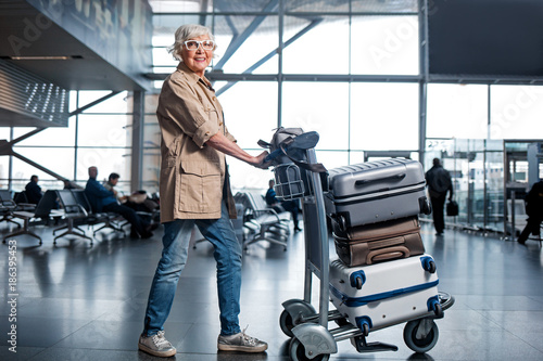 Rest with pleasure. Full length portrait of happy elderly woman is walking along terminal lounge and pushing forward airport trolley with suitcases. She is looking at camera with joy