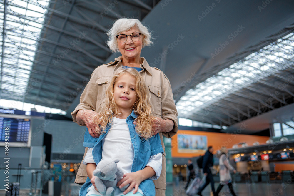 Feeling wonderful. Low angle portrait of cheerful old granny is standing behind her little grandchild and hugging her. They are expressing gladness while waiting for flight at terminal lounge