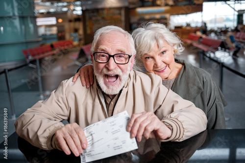 Our happy journey. Top view portrait of aged bearded man and charming old woman are standing together at airport lounge while looking at camera with joy. Gentleman is showing their ticket with smile