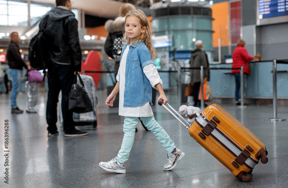 Favorite holiday. Full length portrait of optimistic trendy little girl is walking along airport lounge while carrying orange suitcase. She is looking at camera with slight smile. People in background