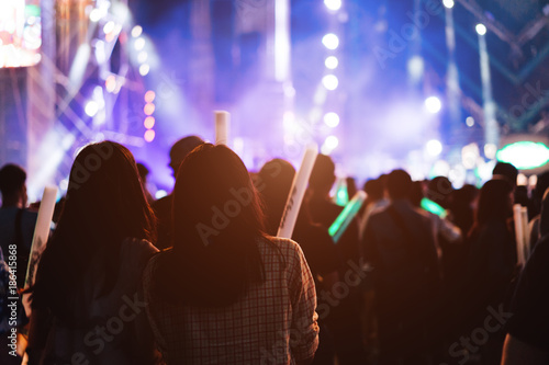 Two women friends crowd concert stage lights and people fan audience silhouette raising hands glow stick in the music festival rear view with spotlight glowing effect