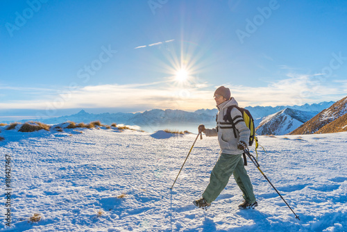 Woman backpacker trekking on snow on the Alps. Rear view, winter lifestyle, cold feeling, sun star in backlight, hiking poles.