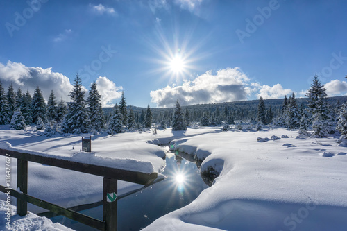 Winter mountain landscape with stream and wooden bridge. Trees and the sun are reflected in the water. Izera Mountains, Czech Republic, Europe.