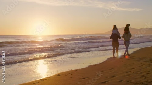 Young family walking and playing on beach with children on shoulders in sunset