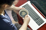 A woman is sitting on the floor cross-legged and working on a laptop. In the hands of a cup of coffee. On the floor is a red plaid.
