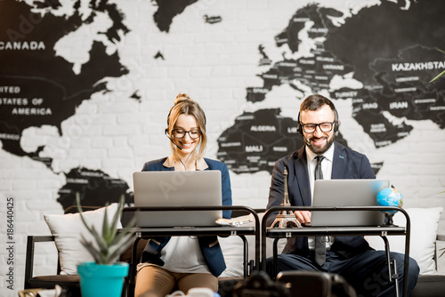 Couple of travel managers working online with laptops and headsets at the agency office with world map on the background