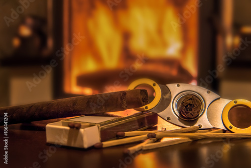Time to enjoy! Quality cigars lie on a wooden table and other accessories such as cigar cutters and matches. In the background a fireplace. Focus on the cigars. Concept: life style or health