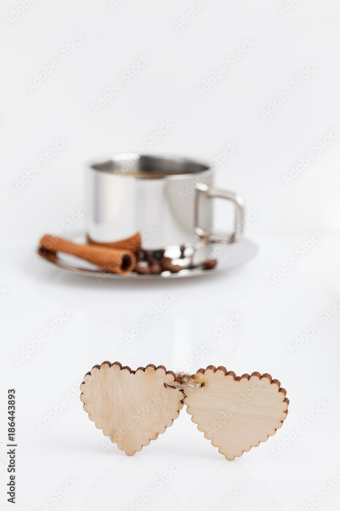 Two wooden hearts connected by a twine on a white table. At the back is a cup of coffee with grains and a cinnamon stick.
