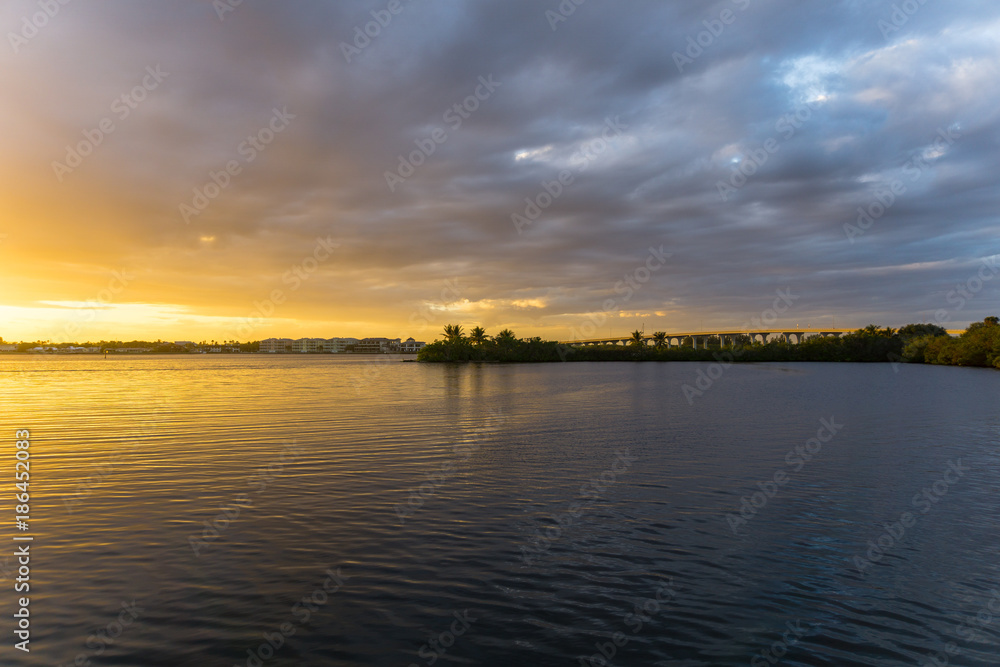 Fototapeta premium USA, Florida, Orange glowing sky with some clouds and highway bridge behind reflecting water