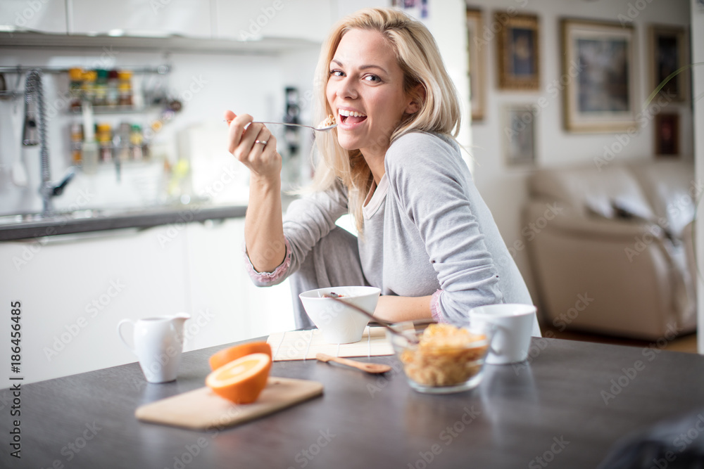 Beautiful blond  caucasian woman posing in her kitchen, while drinking coffee or tea and eating a healthy breakfast meal full of cereal and other healthy foods, including fruit