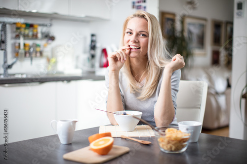 Beautiful blond caucasian woman posing in her kitchen, while drinking coffee or tea and eating a healthy breakfast meal full of cereal and other healthy foods, including fruit