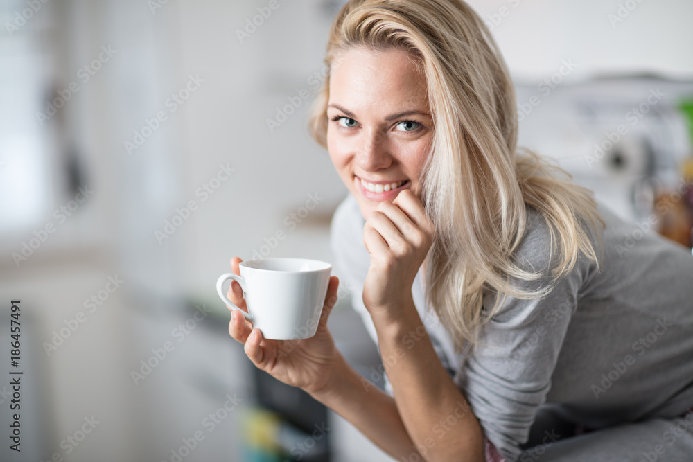 Beautiful blond  caucasian woman posing in her kitchen, while drinking coffee or tea and eating a healthy breakfast meal full of cereal and other healthy foods, including fruit