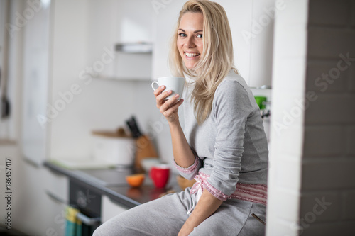 Beautiful blond  caucasian woman posing in her kitchen, while drinking coffee or tea and eating a healthy breakfast meal full of cereal and other healthy foods, including fruit