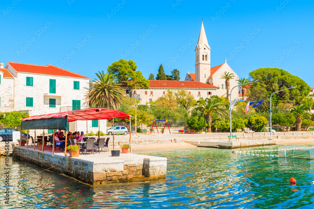 Naklejka premium SUMARTIN PORT, BRAC ISLAND - SEP 13, 2017: people dining in restaurant in Sumartin port with beautiful church in background, Brac island, Croatia.