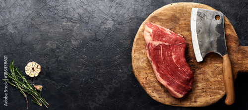 Raw fresh marbled meat Black Angus steak and meat cleaver on wooden board. Meat on black background with rosemarym garlic. Copy space. Top view.