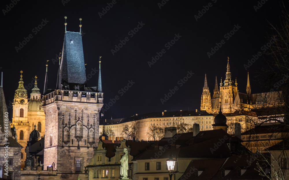 A night view of Prague taken from the Charles bridge depicting multiple towers and spires - the historic skyline and Prague castle on the hill. The historic landmarks are illuminated.