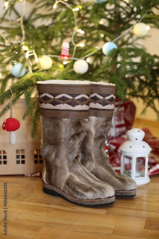 Fototapeta premium a pair of warm boots in wool stands near the Christmas tree with presents at home on a wooden floor. next to a candlelight and a red fly agaric