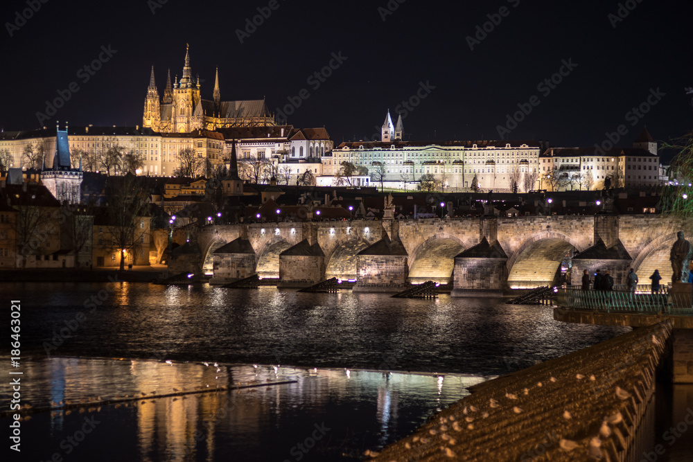 A night photo of Prague with reflections in water. In view Illuminated Charles bridge in foreground, historic skyline of Lesser Side and Prague castle on the hill above the Vltava (Moldau) river.