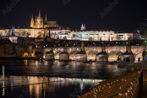 A night photo of Prague with reflections in water. In view Illuminated Charles bridge in foreground, historic skyline of Lesser Side and Prague castle on the hill above the Vltava (Moldau) river.