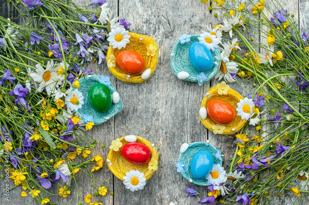 Colorful Easter eggs lying on the wicker plates in the form of a nest on wooden background with a bouquet of wild chamomile flowers and bells. Copy space. Rustic Easter decor.