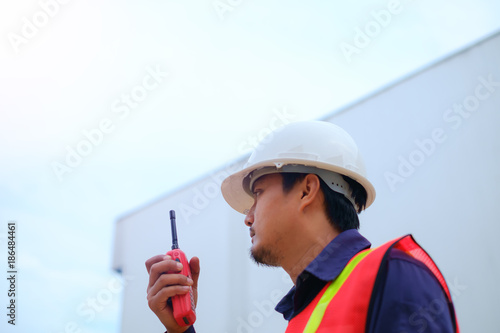 Asian engineer in safety uniform and white helmet using walkie-talkie on blurred industry plant background for industrial concept