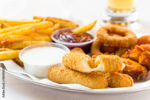 Finger food plate with mozzarella sticks, onion rings, chicken wings, french fries and dip on white table.